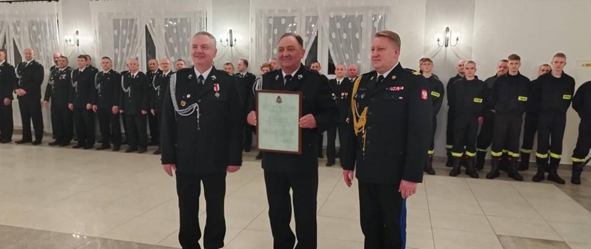 nadbrygadier Grzegorz Janowski oraz dwóch druhów OSP pozują do zdjęcia w czasie uroczystości, druh stojący po środku prezentuje dokument w ramce