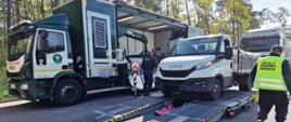Dostawczak skontrolowany przez inspektorów warmińsko-mazurskiej Inspekcji Transportu Drogowego z wykorzystaniem Mobilnej Jednostki Diagnostycznej. W centrum zdjęcia rozstawiona jednostka. Na rozstawionej platformie stoi kontrolowany dostawczak. Za nim ciężarówka. Po prawej tyłem stoi inspektor ITD.
