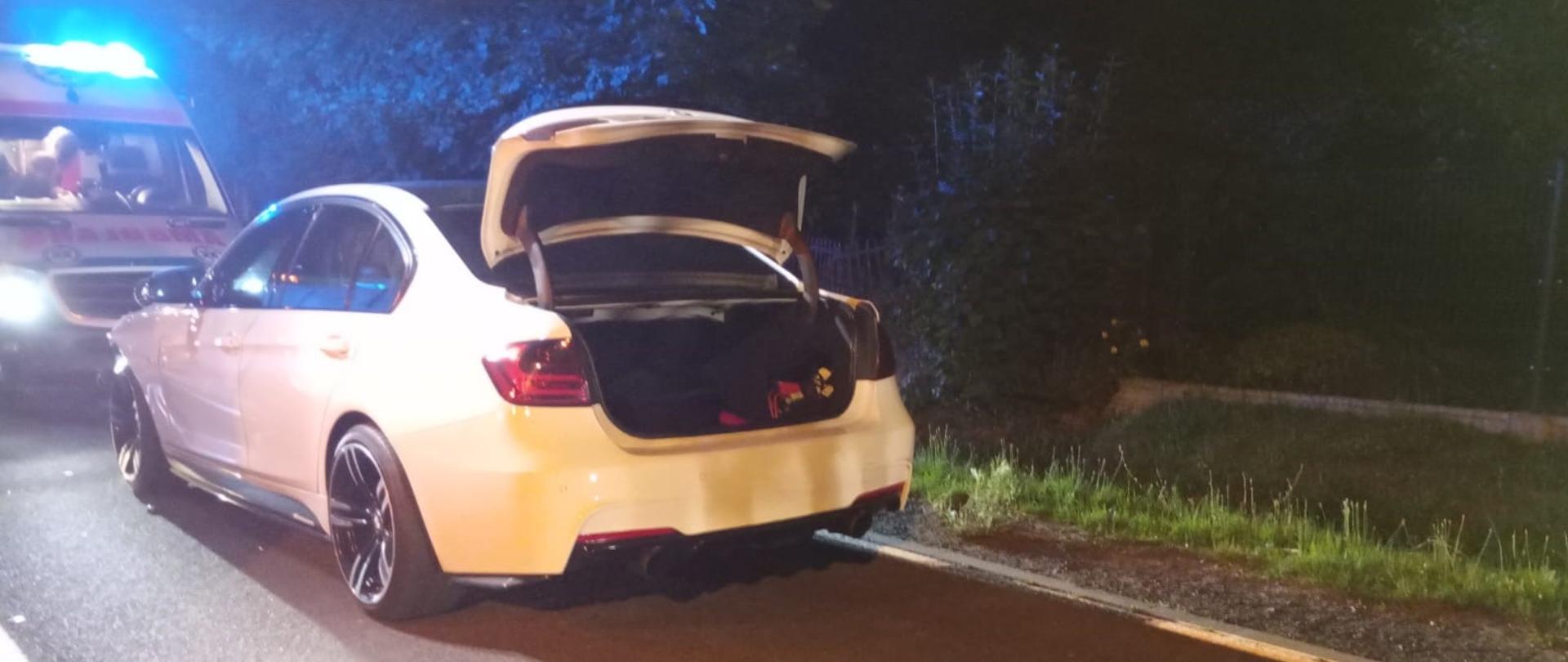 Zdjęcie przedstawia tył białego samochodu osobowego marki BMW, stojącego na poboczu drogi w nocy. Klapa bagażnika jest otwarta, a w środku widać ciemne wnętrze. Obok samochodu, po lewej stronie, stoi karetka pogotowia ratunkowego z włączonymi niebieskimi światłami, które oświetlają otoczenie. Po prawej stronie drogi widoczne jest pobocze porośnięte trawą i krzewami, a dalej ciemne tło drzew. Asfalt jest widoczny, a na nim rzucane są cienie.