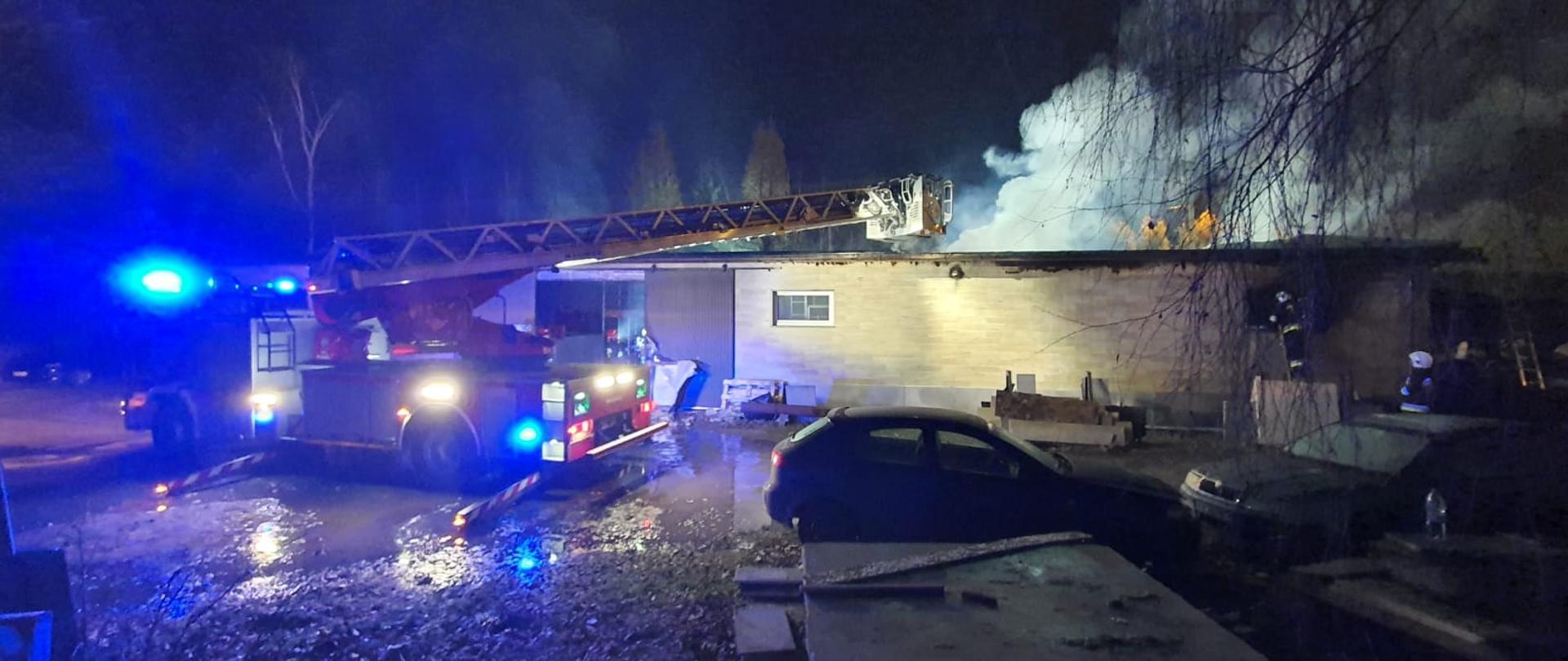 Widok budynku objętego pożarem, 4 ratowników przy drzwiach, sprawiona drabina mechaniczna