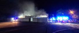 Na zdjęciu w oddali widnieje płonący budynek, który jest otoczony przez zastępy straży pożarnej prowadzące działania ratowniczo-gaśnicze
