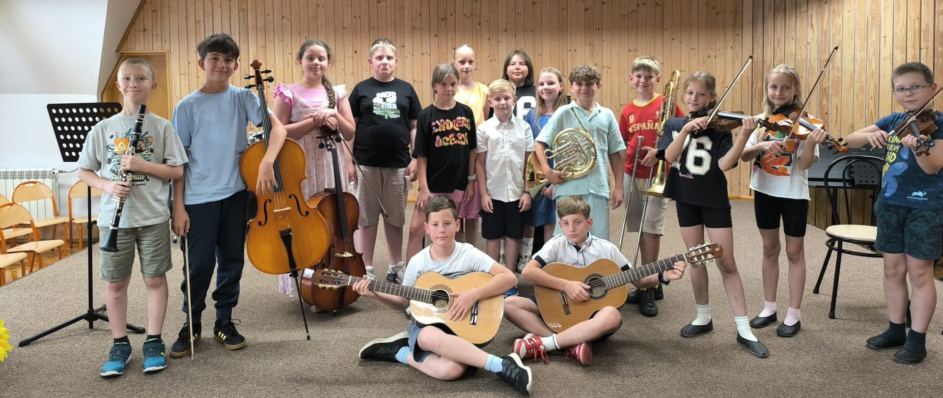 Kilkanaścioro dzieci stoi lub siedzi na scenie ze swoimi instrumentami.