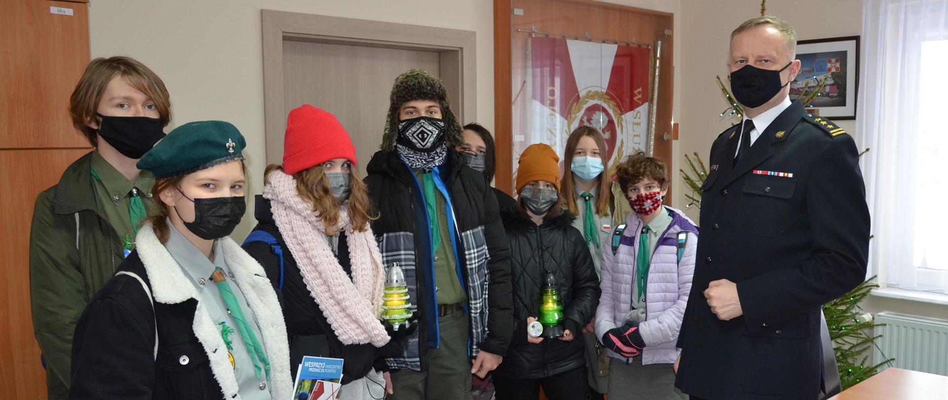 Na zdjęciu widać grupę harcerzy z ZHP Starogard w trakcie przekazania Betlejemskiego Światełka Pokoju. Harcerze trzymają znicze, wszyscy w maseczkach, w tle hoinka
