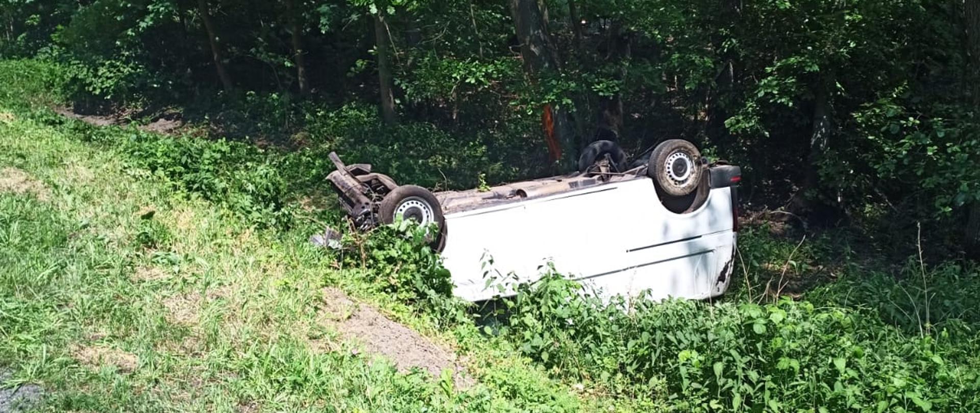 Na zdjęciu samochód dostawczy typu bus leży w rowie kołami do góry.