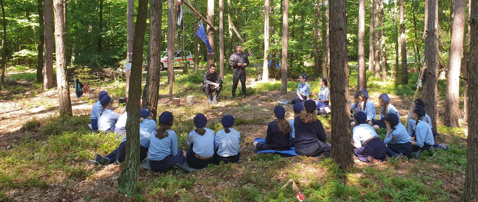 Na zdjęciu widać dwóch funkcjonariuszy w czasie przekazywania informacji harcerkom. Harcerki siedzą na ziemi twarzą do strażaków. Wszystko odbywa się w dużym lesie. 