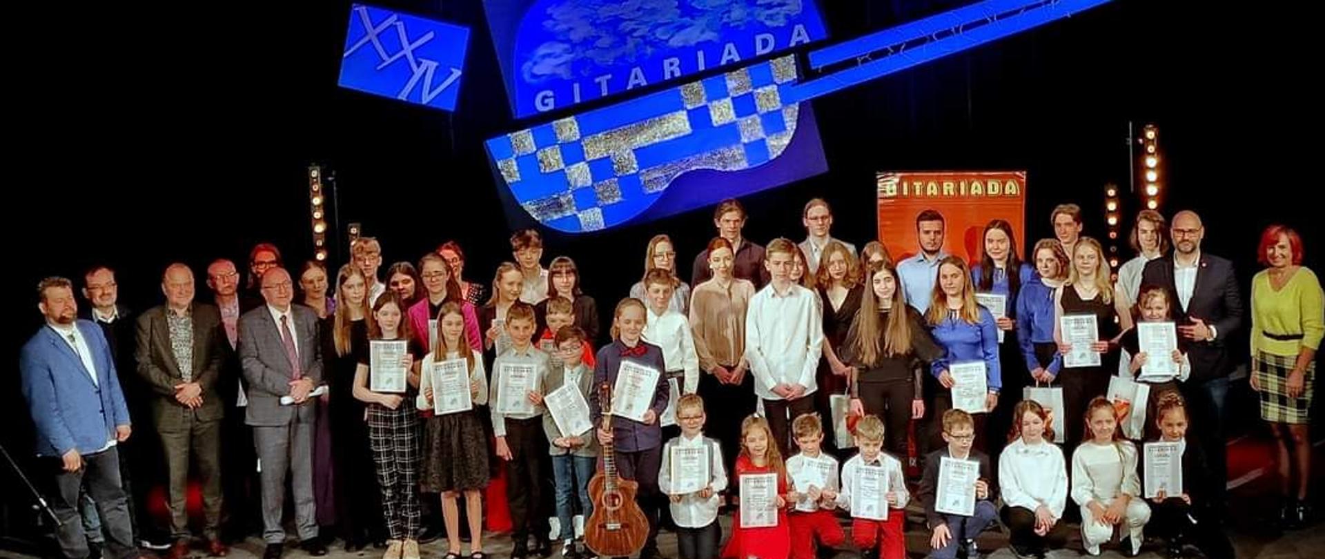 Na zdjęciu wszyscy laureaci XXIV Ogólnopolskiego Konkursu Gitarowego GITARIADA 14-16 kwietnia 2023 trzymający dyplomy oraz nauczyciel W. Kozakowski, Joanna Kocyan, Pani Dyrektor A. Bartkiewicz-Brzozowska, starosta Maciej Awiżeń, burmistrz miasta Kłodzka Michał Piszko, w tle widoczny baner gitary oraz nazwa i data konkursu