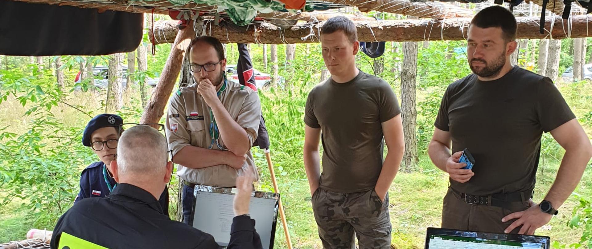 Na zdjęciu tyłem funkcjonariusz Komendy Powiatowej Państwowej Straży Pożarnej w Świeciu przeprowadzający szkolenie z prowadzenia korespondencji radiowej oraz zapoznaje pracowników Lasów Państwowych i uczestników obozu harcerskiego z Wytycznymi Komendanta Głównego Państwowej Straży Pożarnej w zakresie ochrony przeciwpożarowej dla harcerskich obozów namiotowych
