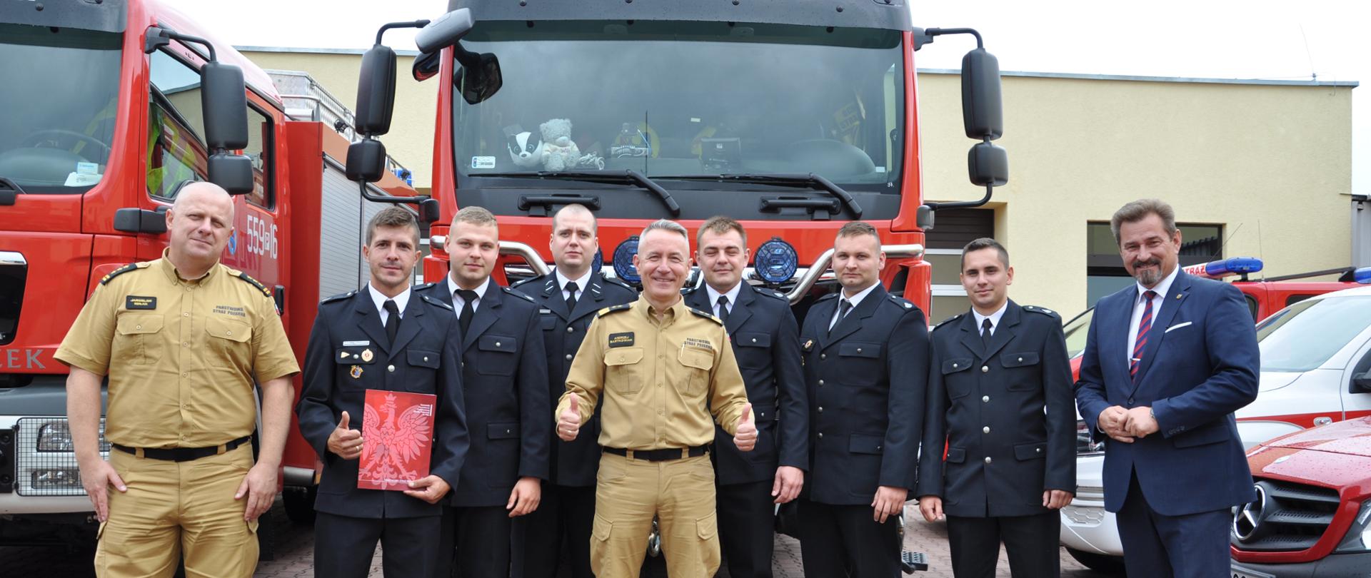 grupa strażaków stoi przed strażackimi samochodami, jeden z nich stojący po środku grupy trzyma dwa kciuki w góre