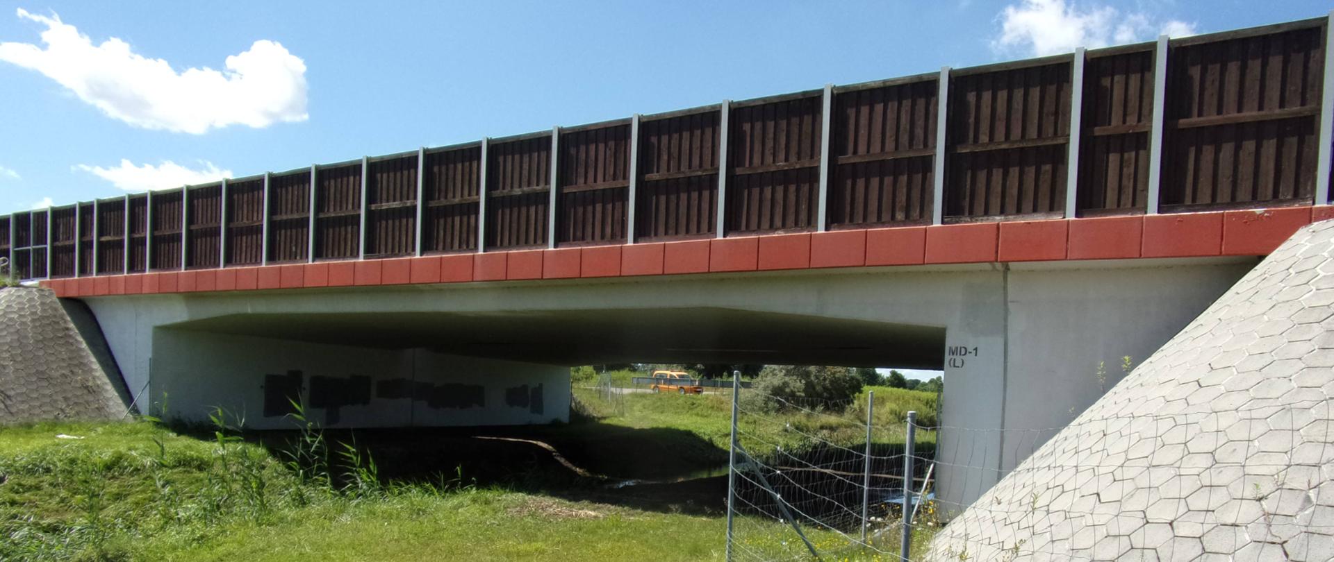 Widok mostu drogowego z umocnionymi skarpami i przestrzenią pod obiektem, z widocznymi elementami konstrukcyjnymi oraz zabezpieczeniem terenu.