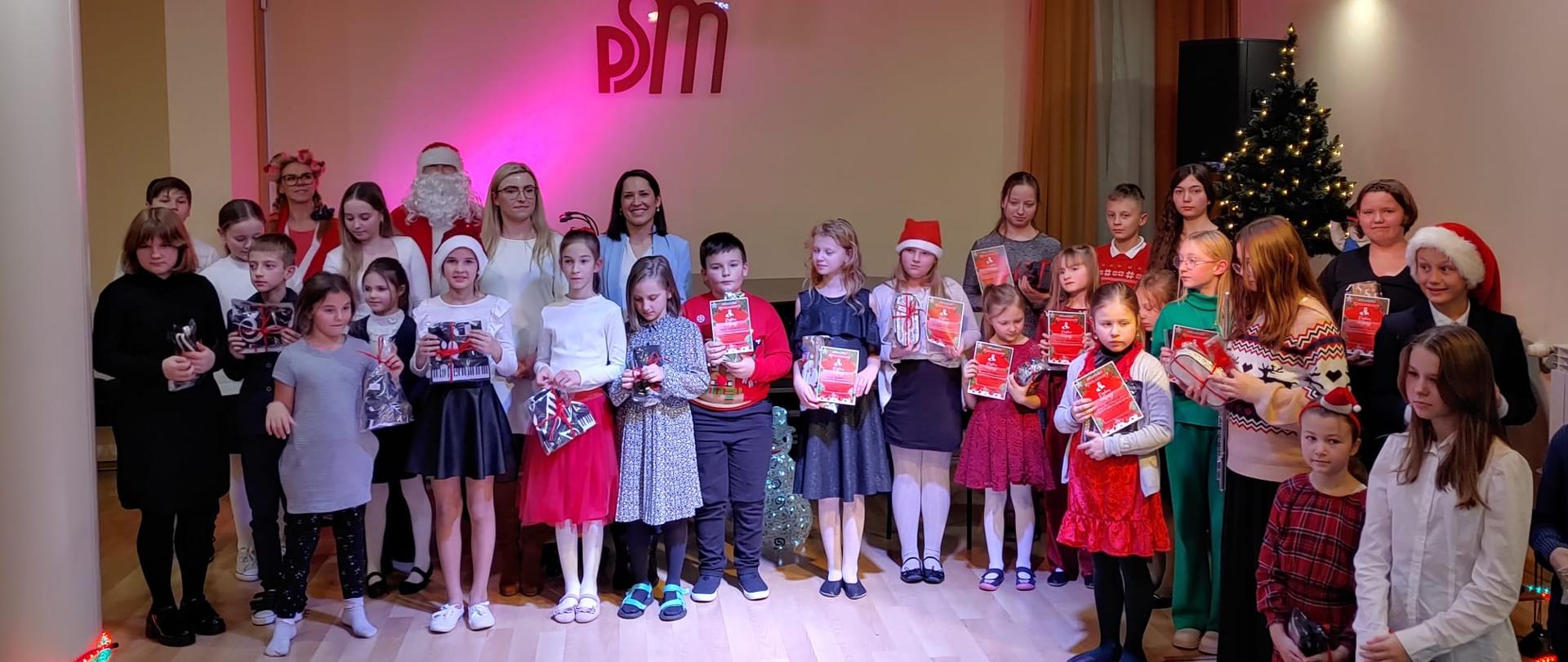 Na zdjęciu widoczne sala koncertowa państwowej szkoły Muzycznej w Starachowicach na środku sceny stoją w 2 rzędach uczniowie którzy brali udział w konkursie na brzegu sceny palą się kolorowe lampki w tle widoczny napis logo PSM Za uczniami stoi Święty Mikołaj już z panią mikołajową oraz członkinie jury