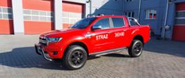 Zdjęcie przedstawia nowy lekki samochód rozpoznawczo - ratowniczy marki FORD. Samochód koloru czerwonego stoi na placu komendy. Z tyłu widać garaże. 