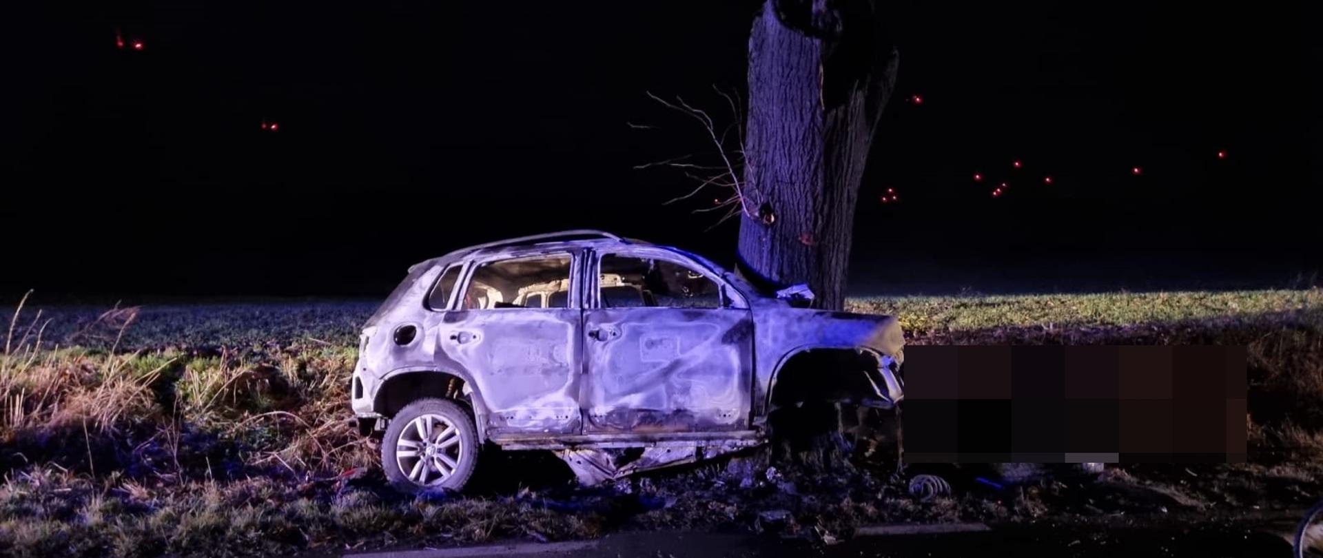 pora nocna, spalony pogięty pojazd po uderzeniu w drzewo. W tle widać pole