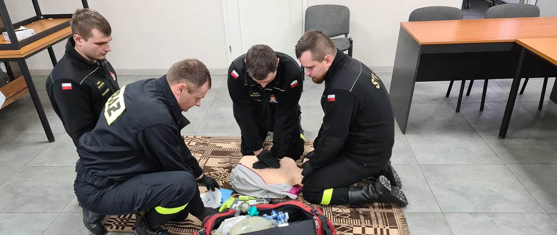 Czterech strażaków wykonuje zadanie egzaminacyjne z zakresu Kwalifikowanej Pierwszej pomocy. Pierwszy wykonuje uciski klatki piersiowej na manekinie, drugi stabilizuje głowę, trzeci i czwarty kompletują sprzęt z torby medycznej. 