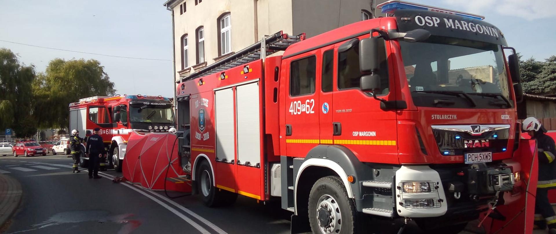 Zdjęcie przedstawia strażaków zabezpieczających pożar mieszkania w budynku wielorodzinnym oraz pojazdy pożarnicze.
W tle budynki wielorodzinne. 
