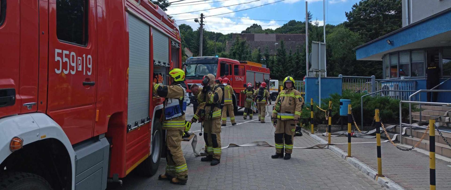 Strażacy i samochody bojowe przed budynkiem administracyjnym.