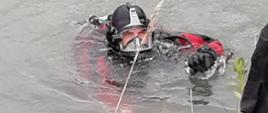 Na zdjęciu widoczny jest nurek PSP znajdujący się w wodzie w czerwonym skafandrze asekurowany z brzegu przy użyciu linek ratowniczych.