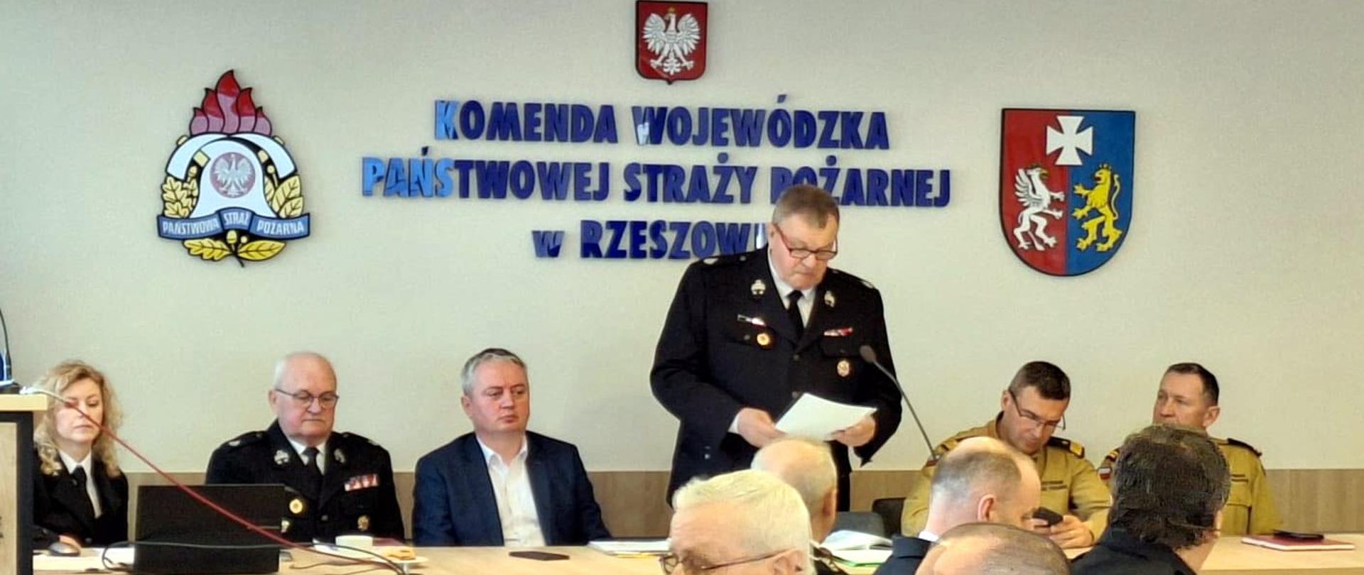 Zdjęcie zrobione podczas spotkania w siedzibie Komendy Wojewódzkiej Państwowej Straży Pożarnej w Rzeszowie. Przy stole prezydialnym siedzi siedem osób, z których jedna przemawia do mikrofonu, czytając z kartki. W tle znajduje się ściana z napisem "KOMENDA WOJEWÓDZKA PAŃSTWOWEJ STRAŻY POŻARNEJ w RZESZOWIE" oraz godło Polski i herb województwa podkarpackiego.