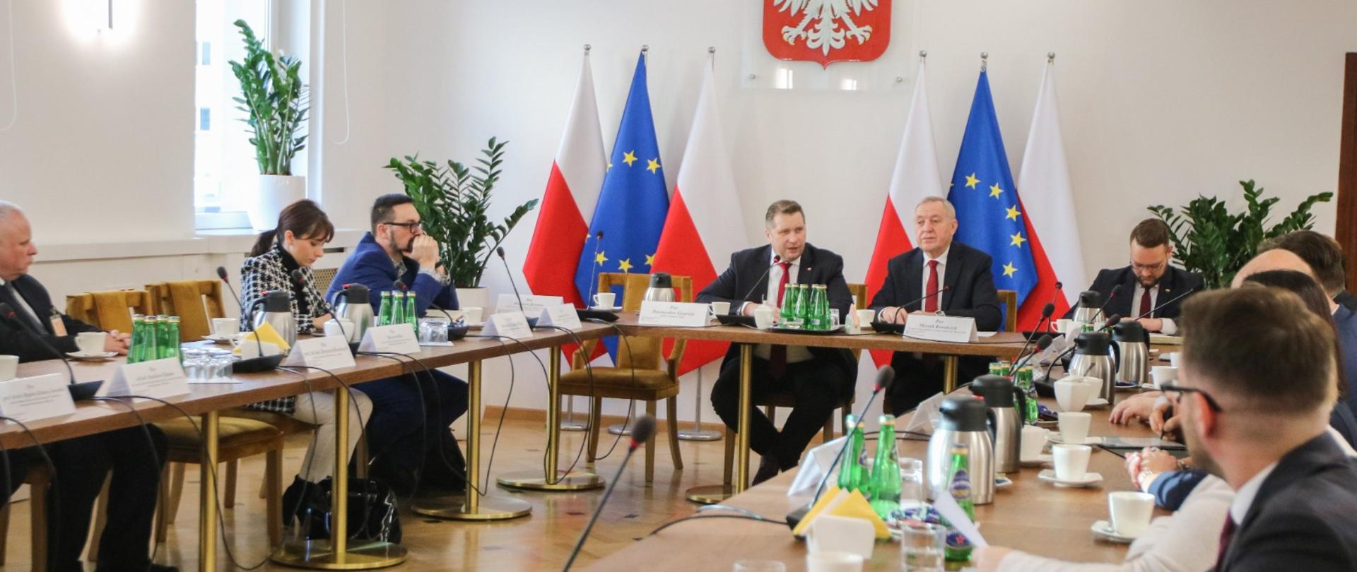 Widok z boku na niewielka salę, przy ustawionych w prostokąt stołach siedzą ludzie, u szczytu stołu siedzi minister Czarnek i mężczyzna w garniturze, za nimi flagi Polski i UE, dalej na ścianie godło.