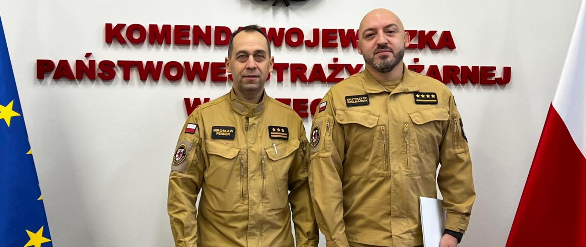 Na zdjęciu stoją dwaj mężczyźni w jasnobrązowych mundurach służbowych. Znajdują się przed ścianą z godłem i napisem instytucji państwowej. Po bokach widać flagę Unii Europejskiej i flagę Polski. Jeden z mężczyzn trzyma białą teczkę. Zdjęcie ma oficjalny charakter.