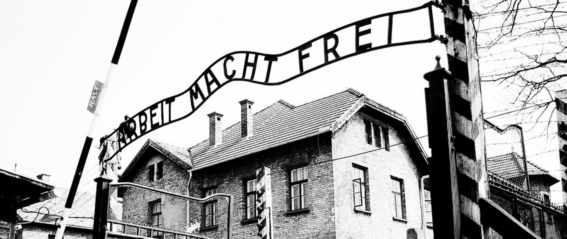Na czarno-białym zdjęciu widoczna na pierwszym planie brama wjazdowa obozu koncentracyjnego Auschwitz z drucianymi wrotami oraz metalowym napisem "Arbeit macht frei". Przed bramą podniesiony szlaban z tabliczką z napisem "halt". W tle za bramą widoczny murowany, jednopiętrowy budynek ze spadzistym dachem.