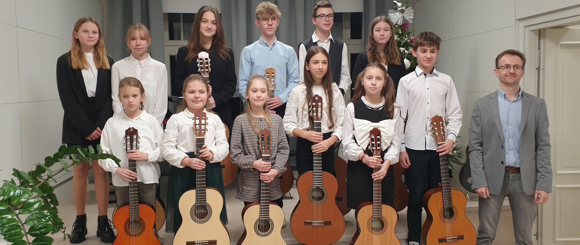Zdjęcie przedstawia uczniów i nauczyciela stojących na scenie w dwurzędzie. Nauczyciel stoi jako pierwszy od prawej strony. Uczniowie trzymają w pionowo gitary. W tle widać rozsunięte kotary w szarym kolorze i okna. Z prawej strony za uczniami widać fragment ubranej choinki, a z lewej zielone gałązki kwiatu. 