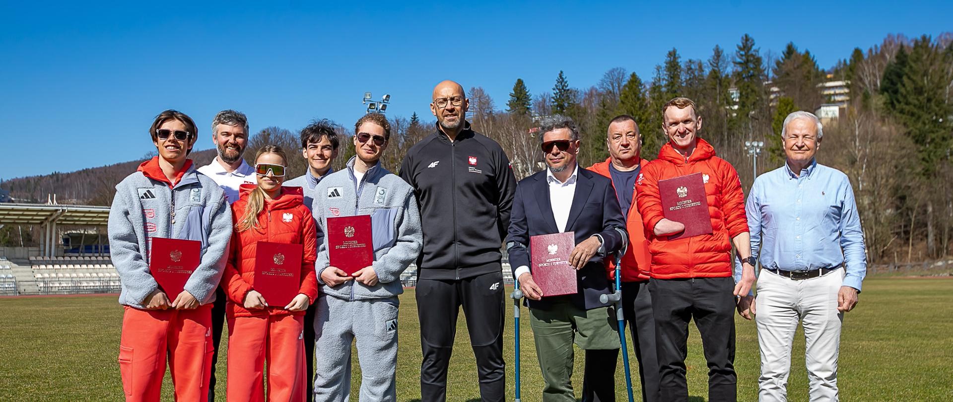 Minister sportu i turystyki Jakub Rutnicki z paralimpijczykami w Wiśle. Stoją w kolorowych dresach na stadionie lekkoatletycznym dla tle zielonego lasu
