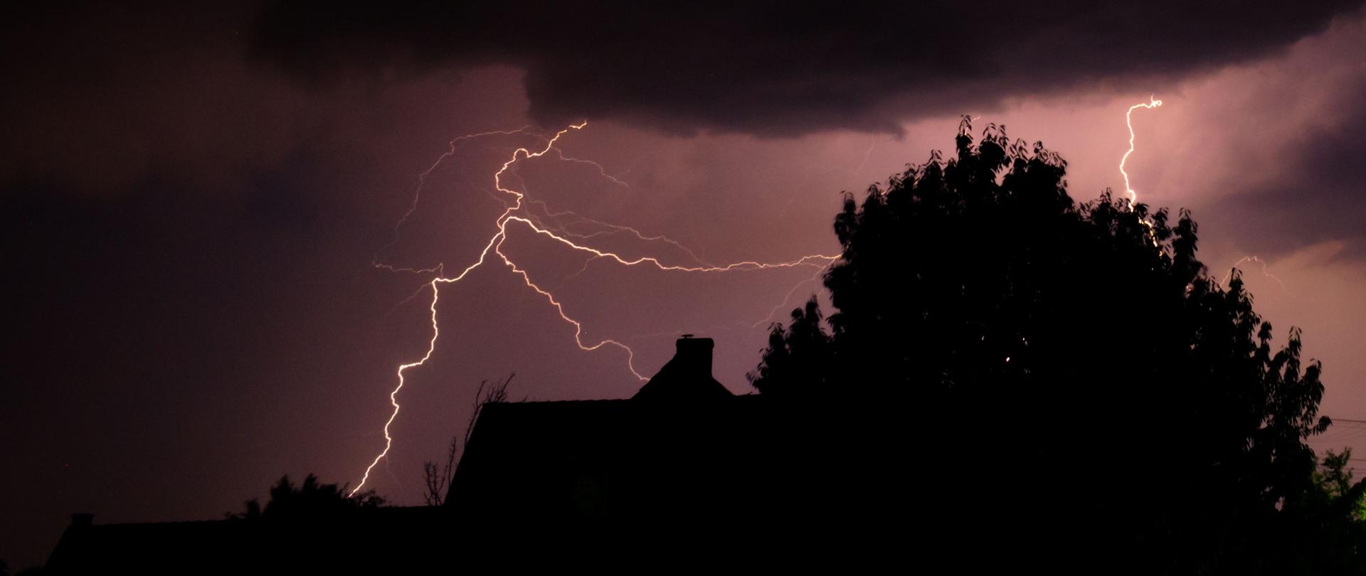 Nocna burza – na ciemnym niebie widoczne rozgałęzione błyskawice oświetlające chmury. Na pierwszym planie zarys drzew i dachów budynków.