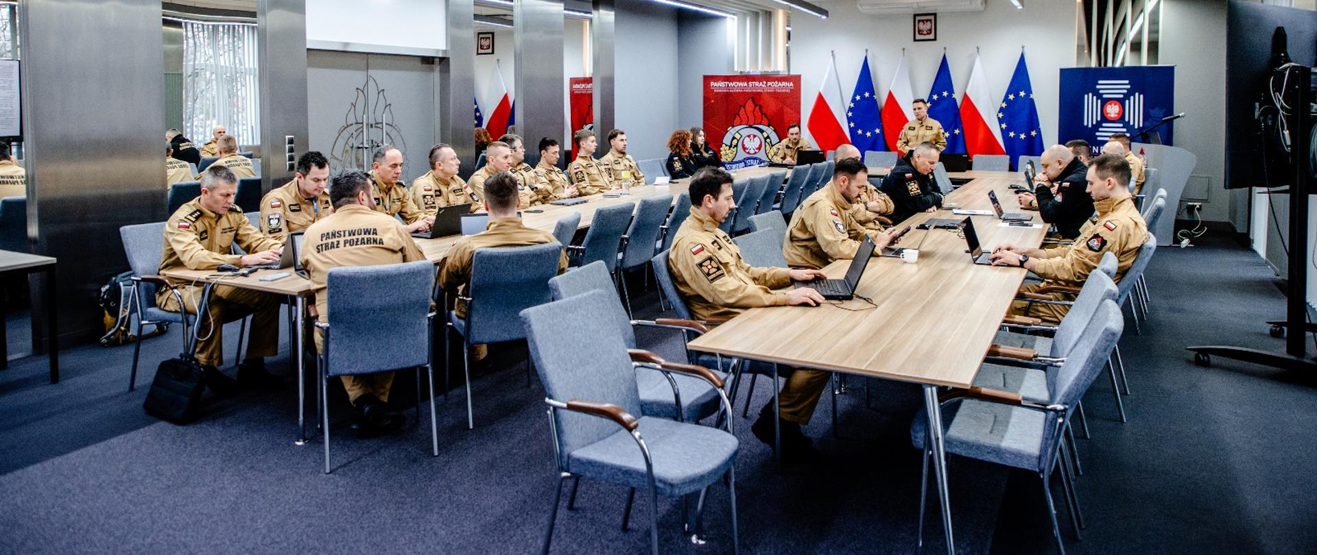Szeroki plan sali konferencyjnej: wielu funkcjonariuszy PSP przy stołach z laptopami; z przodu prelegent, w tle flagi Polski i UE oraz elementy identyfikacji KG PSP