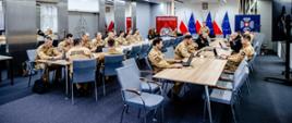 Szeroki plan sali konferencyjnej: wielu funkcjonariuszy PSP przy stołach z laptopami; z przodu prelegent, w tle flagi Polski i UE oraz elementy identyfikacji KG PSP