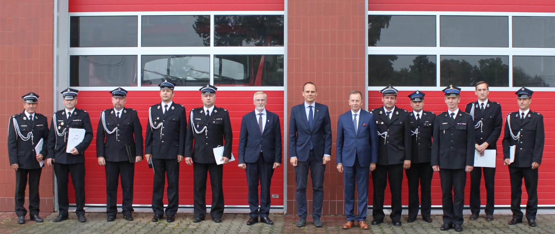 strażacy w mundurach wyjściowych oraz panowie w garniturach na tle czerwonych bram garażowych jednostki ratowniczo gaśniczej