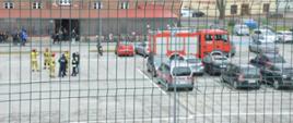 Ćwiczenia w ZSZ. Parking szkolny widziany z góry przez panel ogrodzeniowy. Na parkingu znajdują się strażacy oraz policjanci, a także wozy strażackie i inne zaparkowane pojazdy. W tle wiata, przed którą stoją uczniowie.