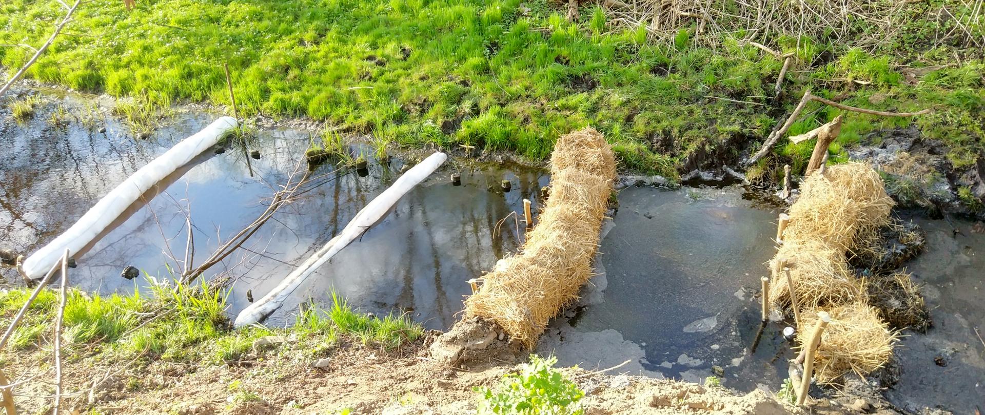Zapory ze słomy oraz zapory sorpcyjne wykonane przez strażaków w korycie rzeki w celu usunięciu plamy nieznanej substancji.