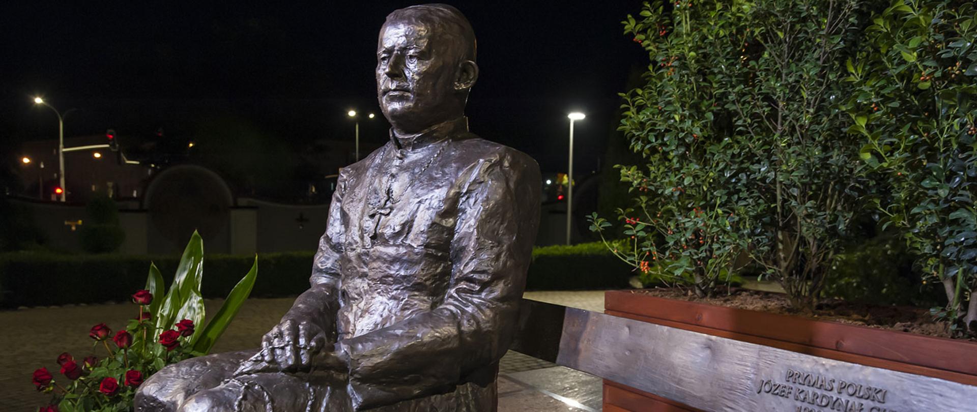 Zdjęcie pomnika Prymasa Polski Józefa kardynała Glempa w formie ławeczki. Józef kardynał Glemp siedzący po lewej stronie ławki. Pomnik odlany z brązy. Zdjęcie wykonane po zachodzie słońca w świetle fleszy.