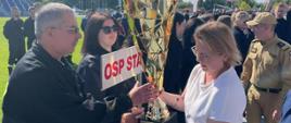 Zdjęcie wykonane w Radomiu na stadionie Miejskiego Ośrodka Sportu i Rekreacji podczas rozgrywek Zawodów Sportowo – Pożarnicze Ochotniczych Straży Pożarnych o Puchar Minister Klimatu i Środowiska. Na zdjęciu widać ustawionych w dwu szeregu członków jednej z drużyn Ochotniczych Straży Pożarnych biorących udział w zawodach. Przed nimi idą trzymając puchar w rękach minister klimatu i środowiska pani Anna Moskwa w asyście komendanta głównego Państwowej Straży Pożarnej generała brygadiera Andrzeja Bartkowiaka. Za chwilę pani minister dokona wręczenia pucharu nagrody zwycięskiej drużynie. 