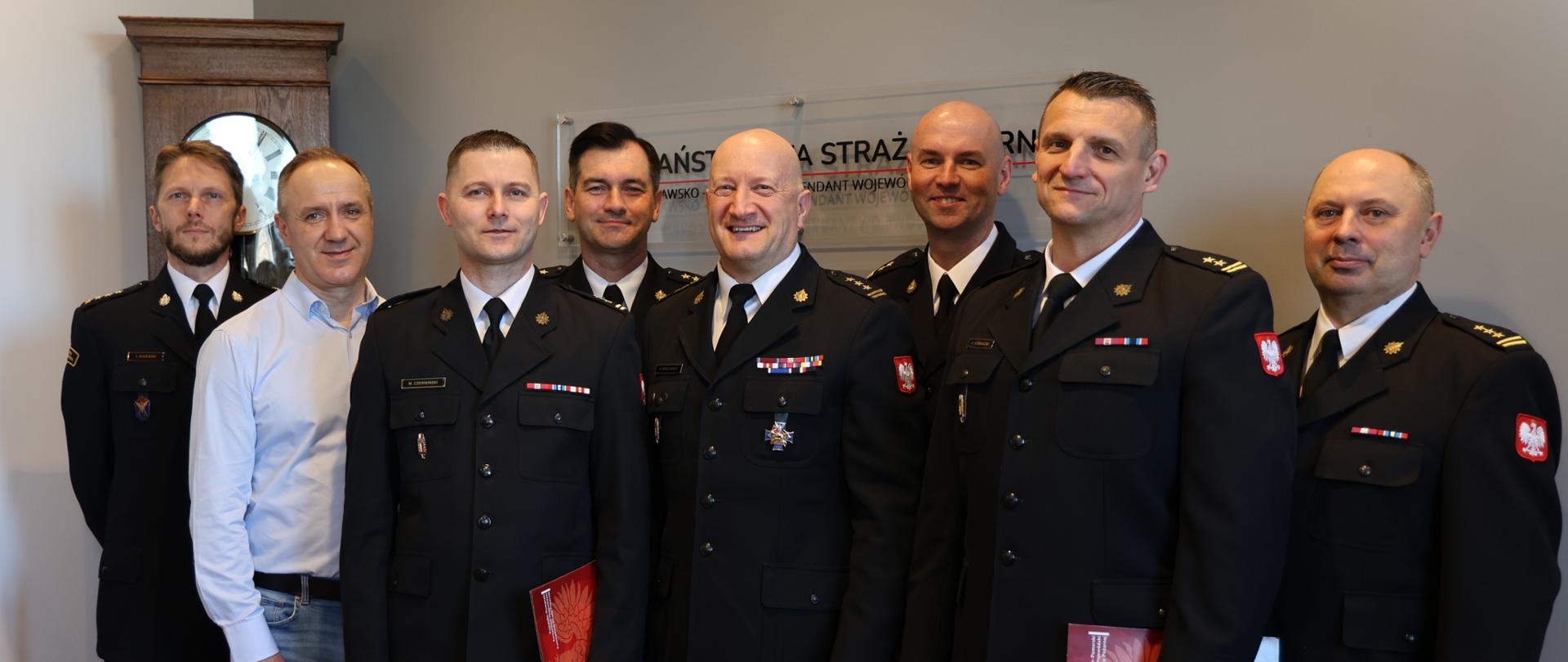 Siedmiu strażaków w mundurach wyjściowych oraz jeden mężczyzna w błękitnej koszuli stoją w grupie w gabinecie komendanta. Dwóch z nich trzyma w ręku czerwone teczki na dokumenty. Za nimi stojący zegar i napis firmowy na ścianie. 