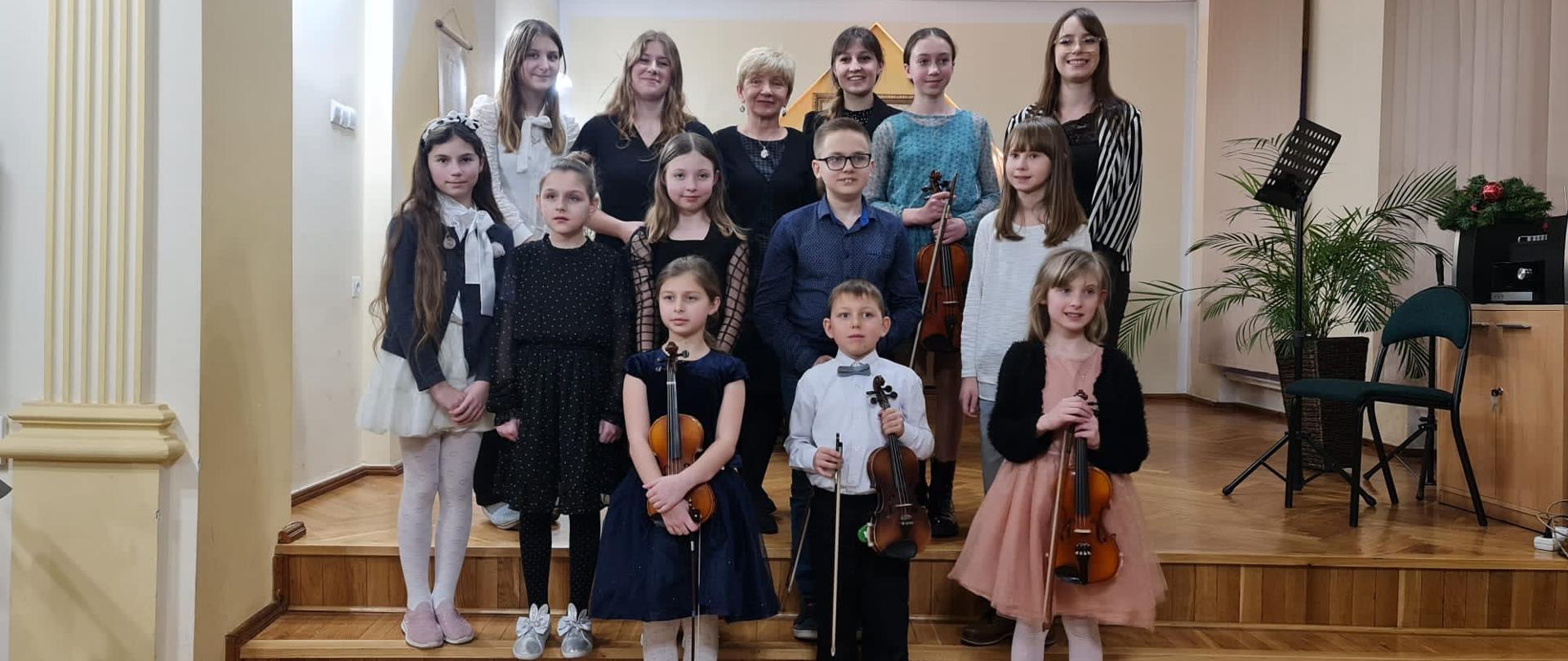 Zdjęcie grupowe uczniów występujących na koncercie klasy skrzypiec P. Karoliny Lizurej i P. Natalii Mierzwy - 25.01.2023. Na zdjęciu widoczna scena auli PSM I st. w Kłodzku.
