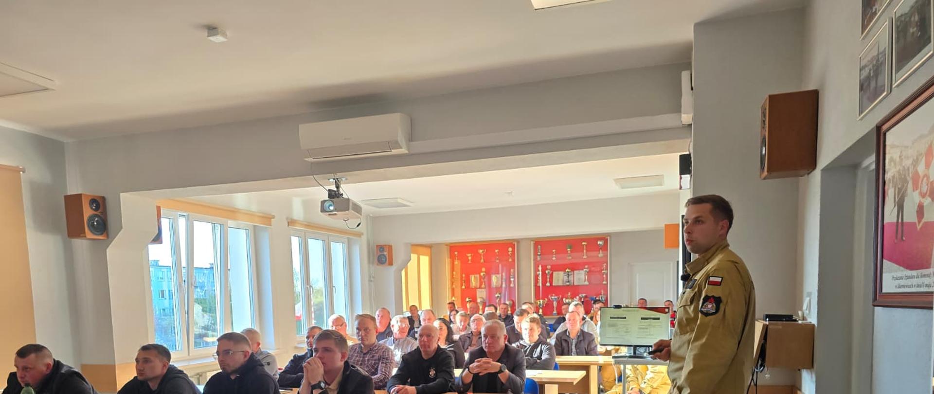 „Funkcjonariusz PSP prowadzi prezentację przed grupą uczestników siedzących przy stołach podczas odprawy służbowej.”