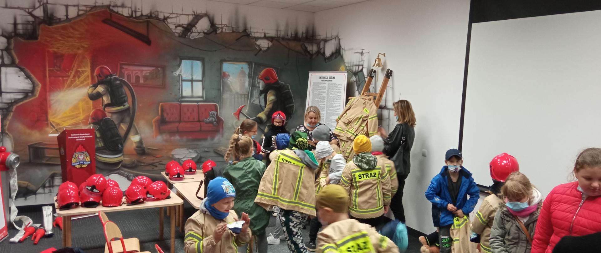Zdjęcie przedstawia dzieci i wychowawców w sali edukacyjnej, która ma na celu poprawę świadomości z zakresu bezpieczeństwa dzieci w wieku przedszkolnym oraz w okresie szkolnym. Dzieci ubierają się w odzież strażacką i hełmy