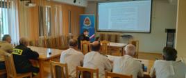 widoczni uczestnicy spotkania, druhowie OSP, oraz funkcjonariusz psp przedstawiający prezentację.