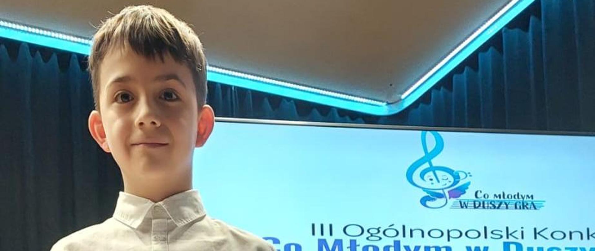 Chłopiec stoi trzymając przed sobą dyplom wyróżnienia, w tle baner z plakatem III Ogólnopolskiego Konkursu "Co młodym d duszy gra?"