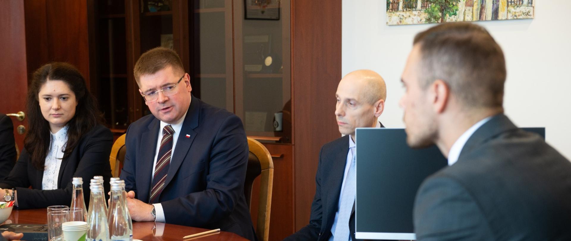 Minister w otoczeniu kilku osób siedzi przy stole, rozmawiają.