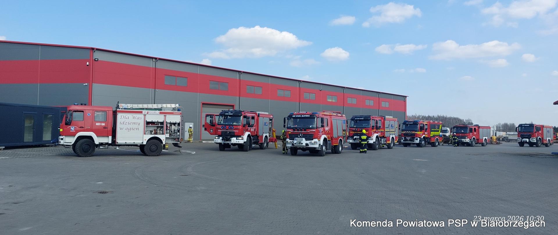 Panorama placu z siedmioma różnymi wozami strażackimi (OSP i PSP) ustawionymi w rzędzie na tle długiej hali. Na jednym z aut widnieje napis: „Dla Was idziemy w ogień”.