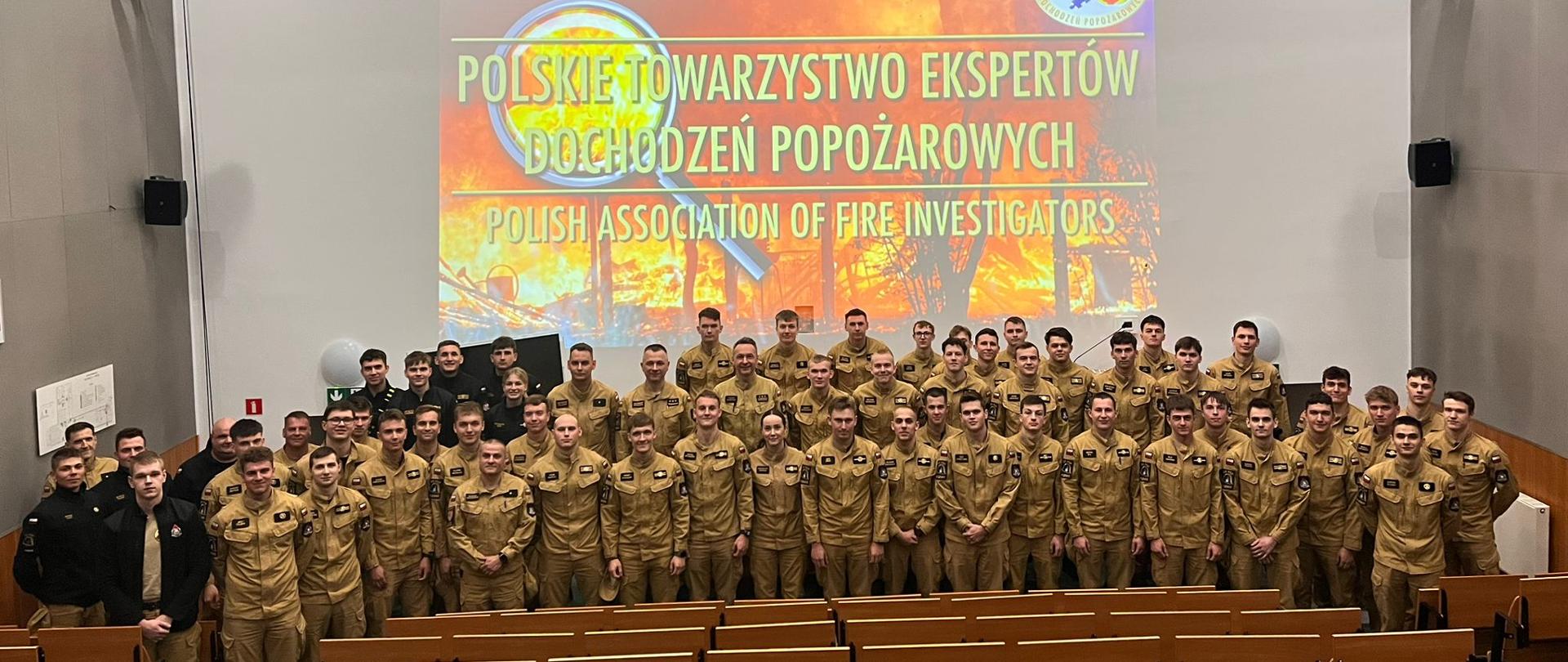 Strażacy stojąc przed katedrą na sali wykładowej rozmawiają z uczestnikami spotkania na auli, w tle wyświetlana jest prezentacja na ścianie.