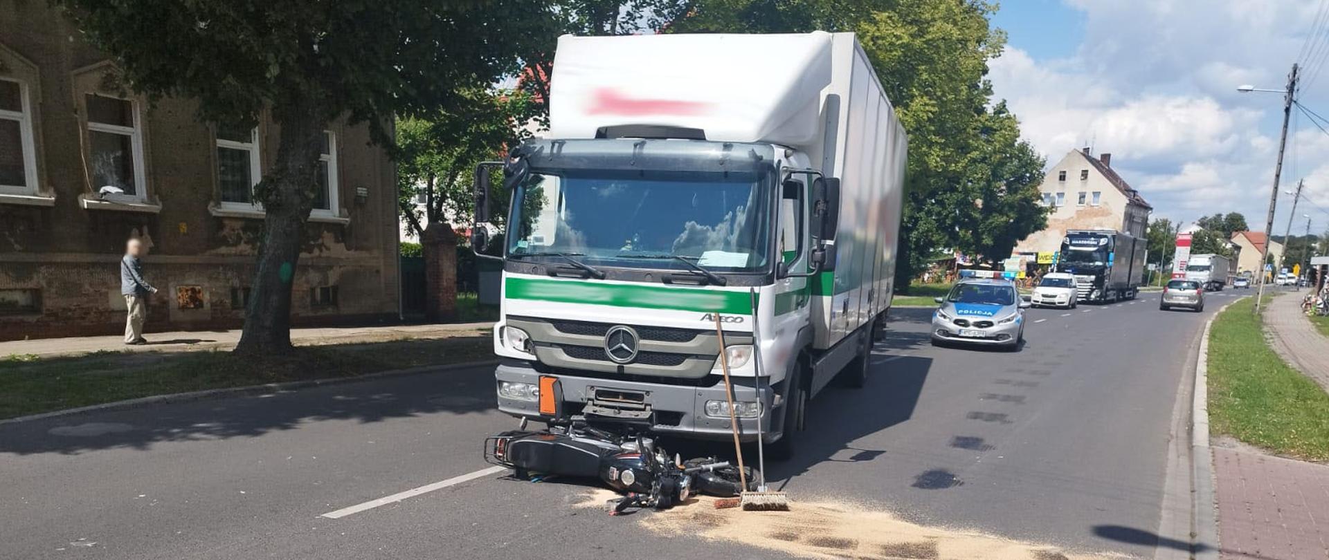 Zdjęcie przedstawia stający na pasie drogowym pojazd dostawczy, pod którym znajduje się motocykl.