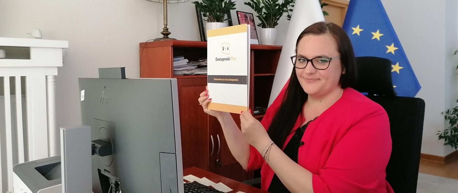Zdjęcie minister w swoim gabinecie. Małgorzata Jarosińska-Jedynak siedzi uśmiechnięta przed komputerem, w dłoniach trzyma teczkę z napisem Dostępność Plus.