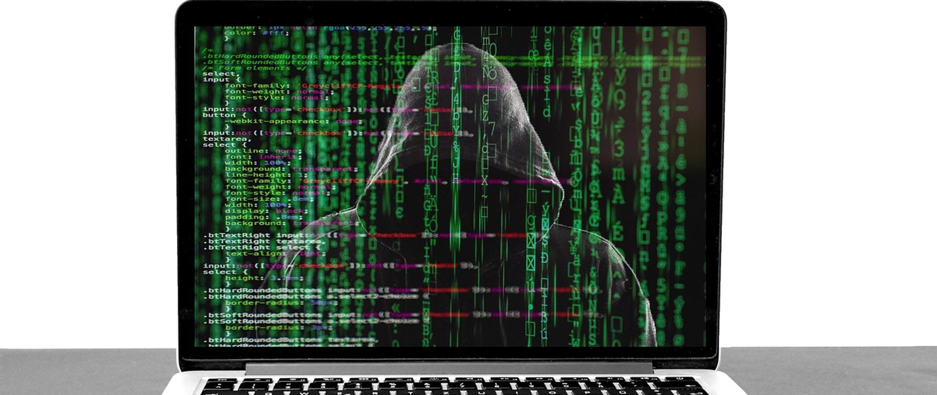 laptop, na ekranie laptopa widać postać w kapturze i dużo zielonych cyferek i literek porozrzucanych na całym ekranie
