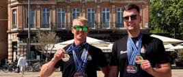 Dwóch mężczyzn stoi na zewnątrz przed dużym banerem informującym, że Albi zostało Europejskim Miastem Sportu 2026. Obaj mają na sobie czarne koszulki i okulary przeciwsłoneczne, a na szyjach liczne medale. Za nimi widoczny jest jasny budynek z dużymi oknami. Zdjęcie ukazuje strażaków z Opolszczyzny po udanych startach w konkurencjach pływackich.
