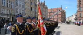 Zdjęcie z uroczystości, strażacy ustawieni w szeregu, odświętnie ubrani, poczty sztandarowe i flagi państwowe w tle.