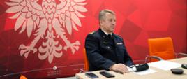 Zdjęcie przedstawia mazowieckiego komendanta wojewódzkiego PSP, który uczestniczy w wideokonferencji poświęconej przygotowaniu do sezonu zimowego. Na pierwszym planie widać komendanta mazowieckich strażaków, który siedzi na krześle. W głębi godło RP biały orzeł w koronie na czerwonym tle.