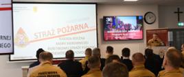 Strażak stoi przy mównicy i przemawia do funkcjonariuszy którzy siedzą na krzesłach na ekranach wyświetlana jest prezentacja.
